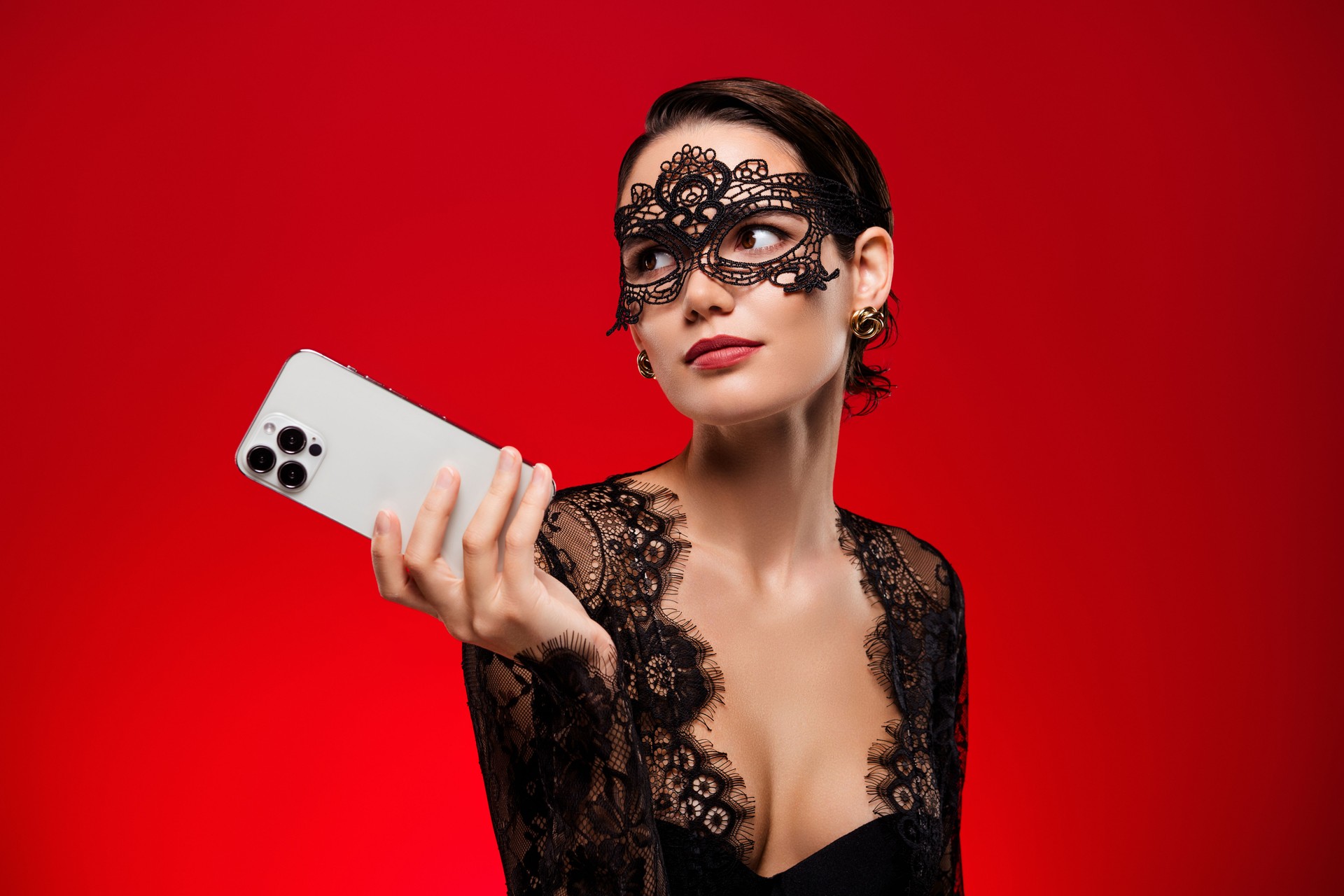 Photo of sexy young girl hold device look empty space dressed black dress mask halloween vip party red color background