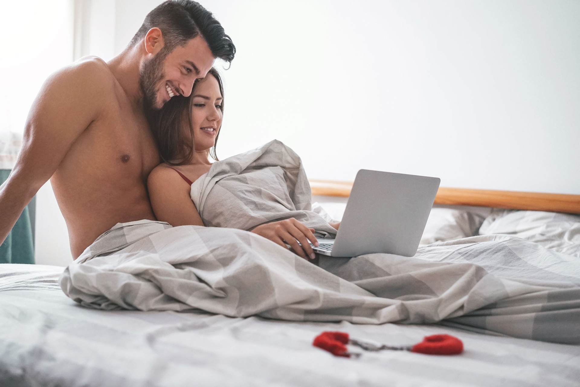Happy couple watching on computer while lying on bed under blanket - Young lover watching role games sex video on laptop - People, sexual, intimate, youth and technology concept